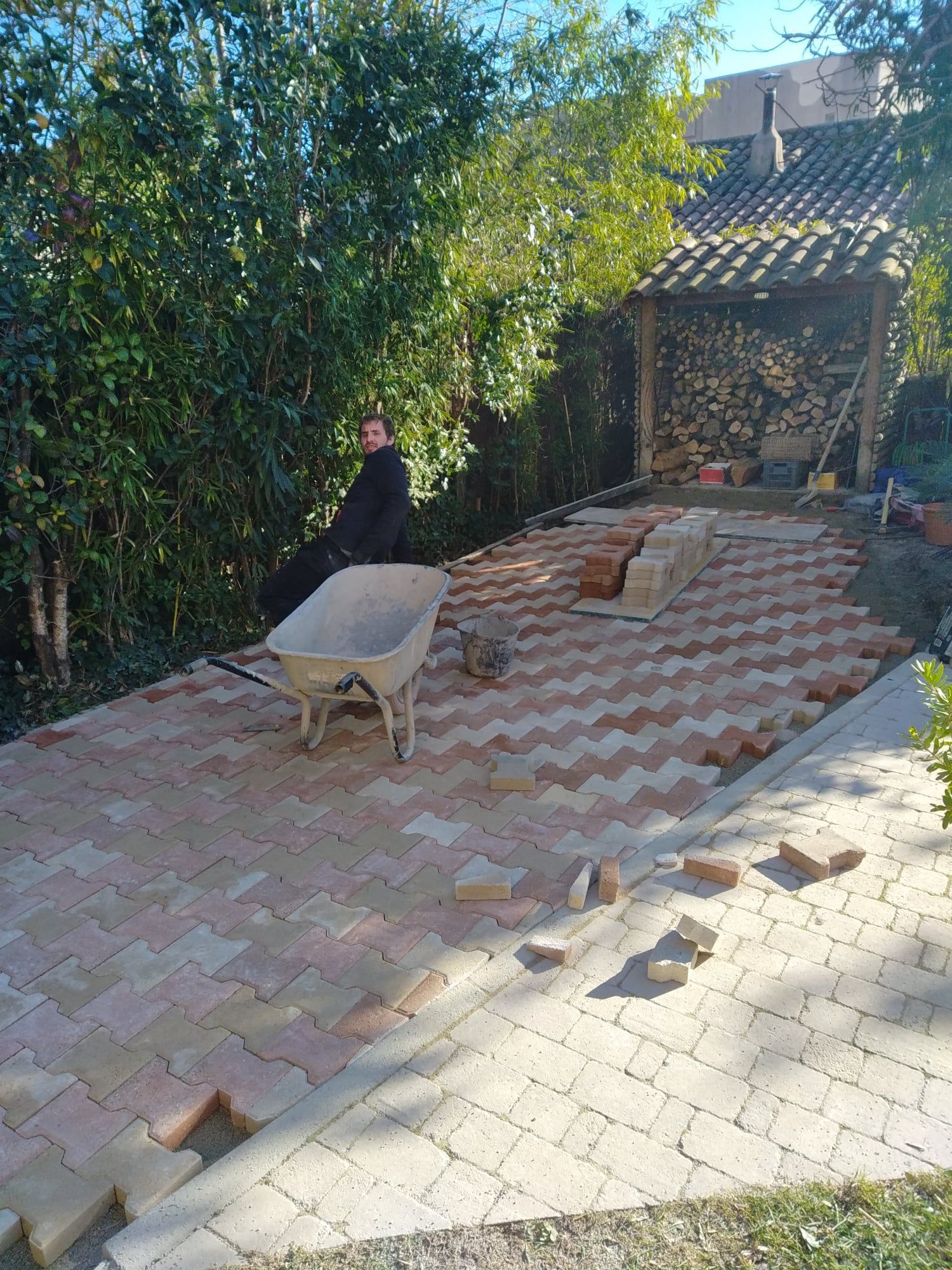 Un homme pose un chemin en briques à chevrons dans une cour arrière ensoleillée avec une brouette.