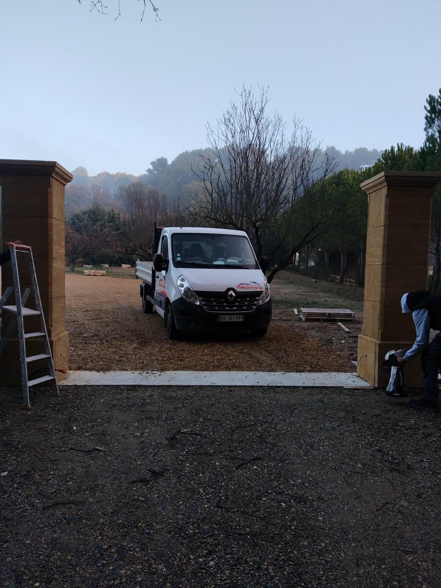 Camion garé entre des piliers de pierre ; ouvriers près de l'entrée du portail sur du gravier, temps brumeux à l'extérieur.