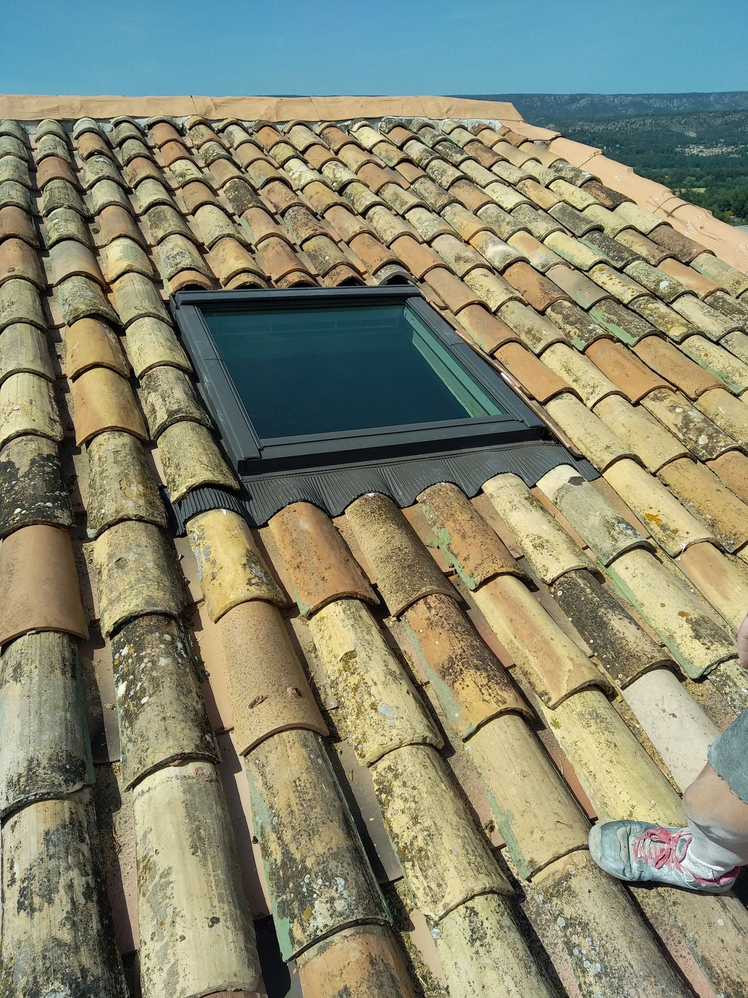 Toiture en ardoise avec deux puits de lumière reflétant un ciel bleu, à côté d'un bâtiment beige.