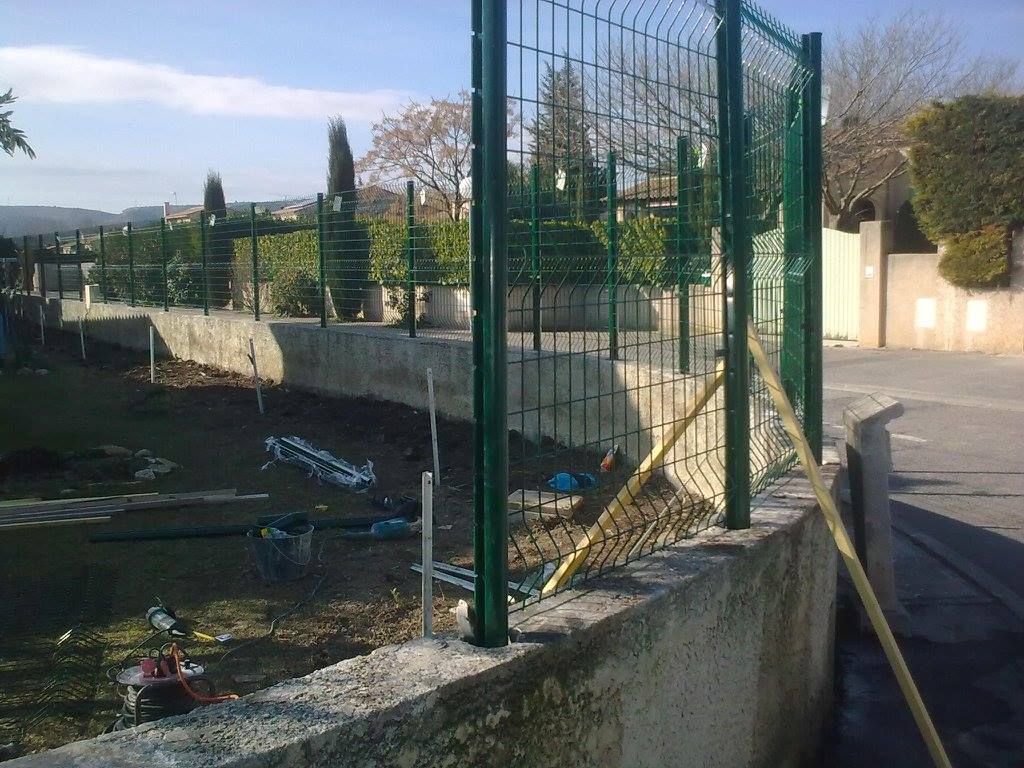 Clôture métallique verte au sommet d'un mur en béton, à côté d'une route, surplombant une zone herbeuse avec des arbres et des bâtiments en arrière-plan.