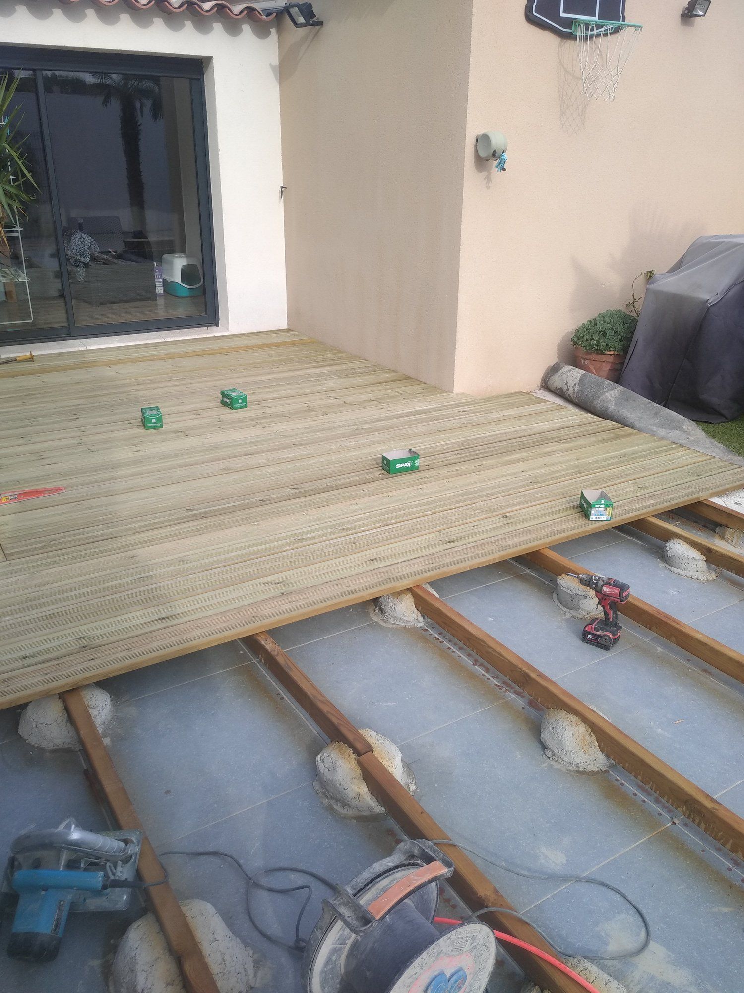 Construction d'une terrasse en bois dans un jardin. Des planches sont posées sur une structure posée sur un socle en béton, avec des outils dispersés un peu partout.