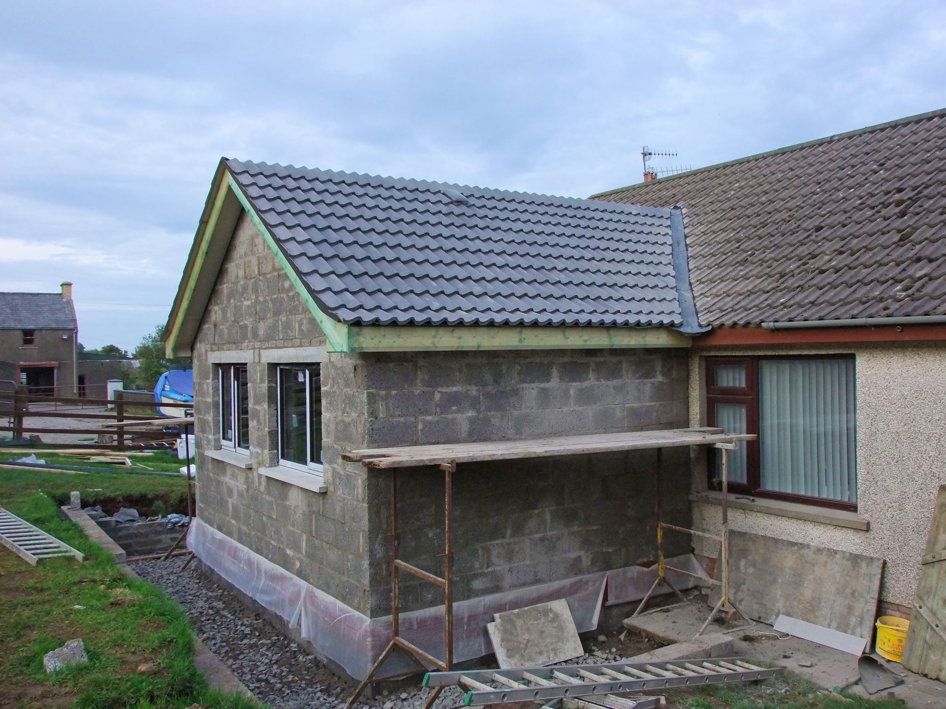 Une extension de maison en construction, murs en blocs gris, toit en tuiles grises, à côté d'une maison aux fenêtres marron, à l'extérieur.
