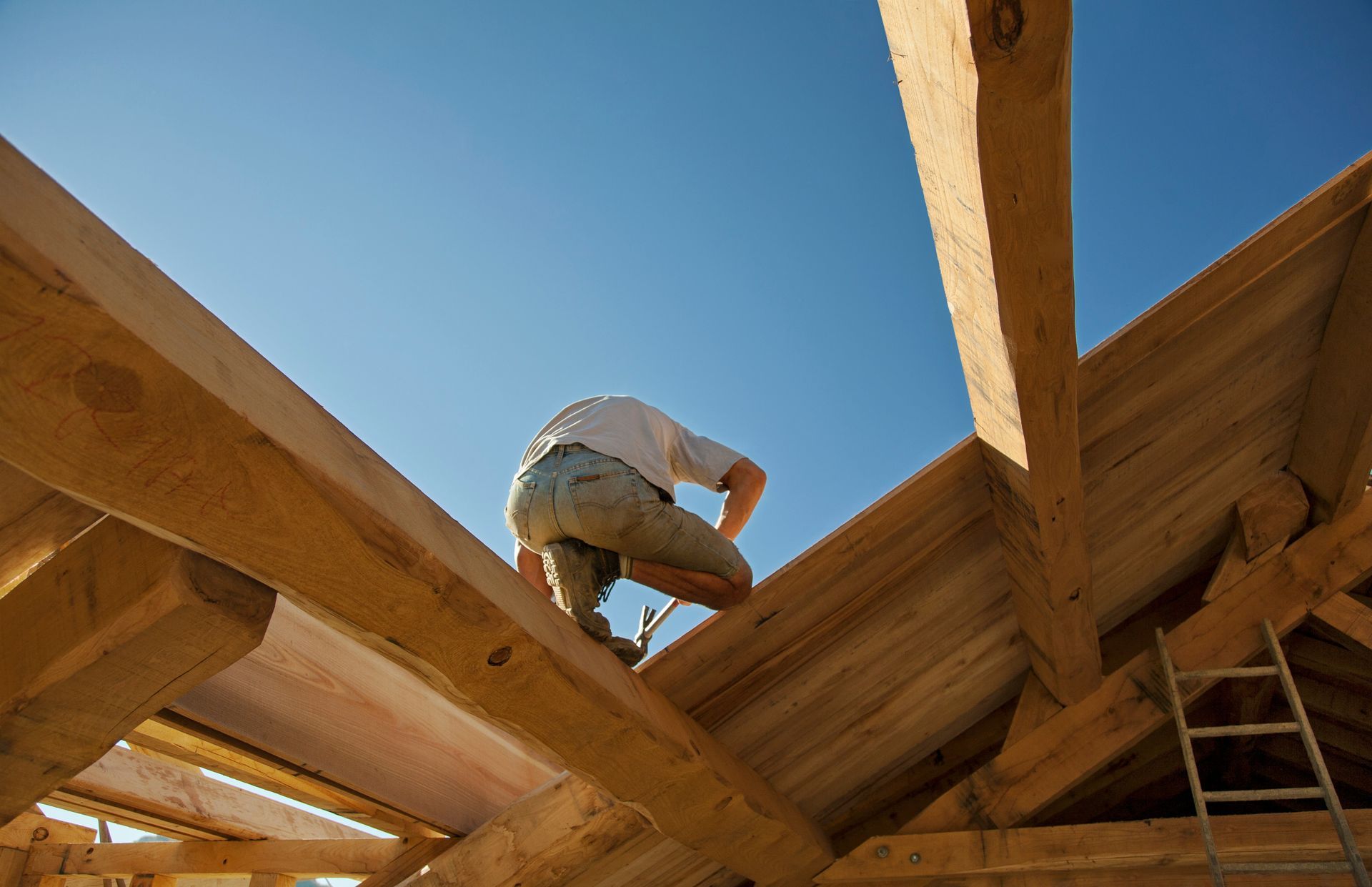 Charpentier travaillant sur une charpente en bois sous un ciel bleu clair.