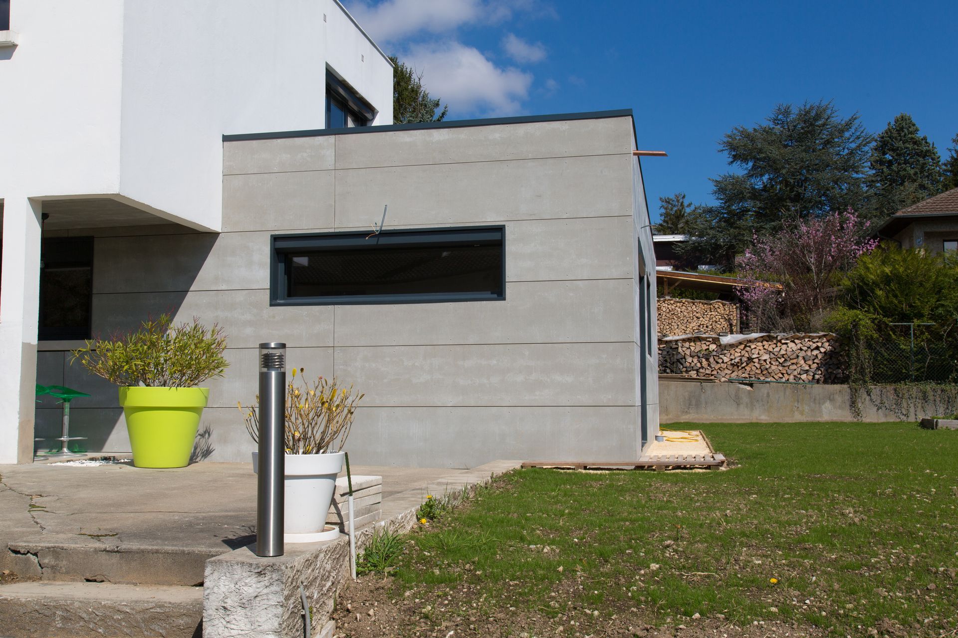 Ajout d'un bâtiment moderne en béton gris avec une petite fenêtre, à côté de la maison blanche et de la pelouse verte.