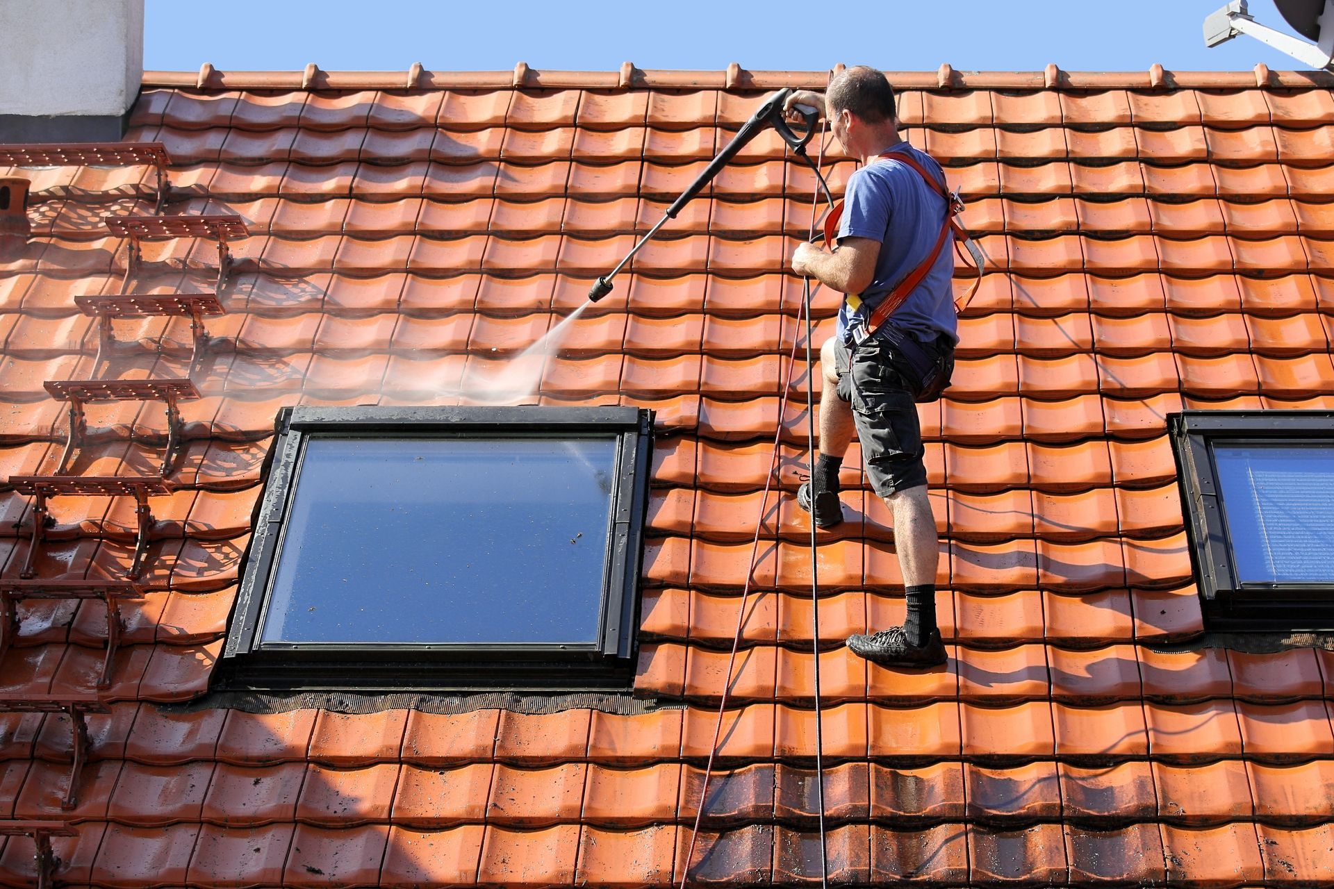 Un homme sur un toit utilise un nettoyeur haute pression pour nettoyer les tuiles près d'un puits de lumière par une journée ensoleillée.