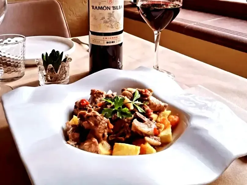 Guiso con patatas y champiñones en cuenco blanco, vino y botella de vino en la mesa.