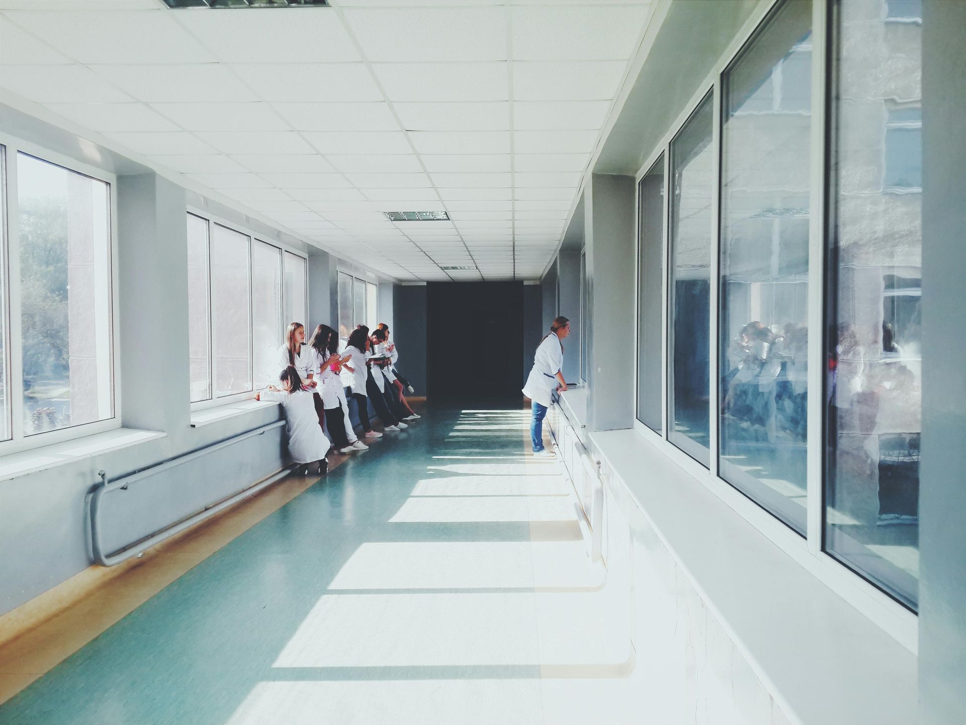 Pasillo del hospital con enfermeras con uniformes blancos mirando por grandes ventanas.