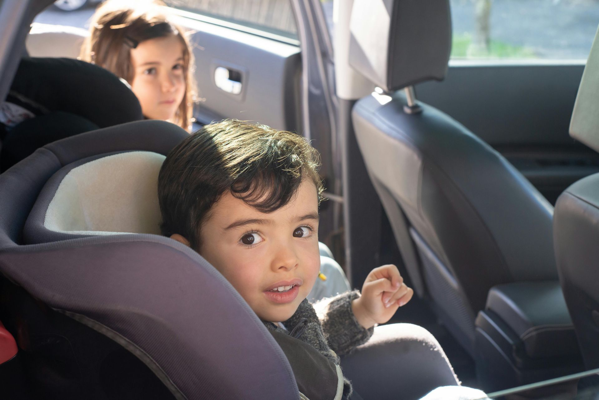 Un niño en el asiento del coche sonríe, mirando a la cámara; una niña al fondo.
