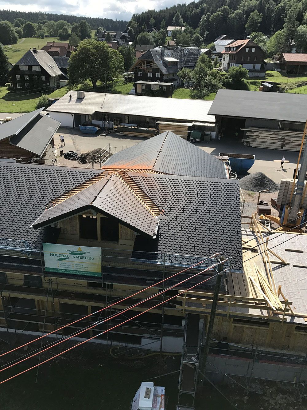 Eine erhöhte Ansicht zeigt ein im Bau befindliches Gebäude mit freiliegenden Dachsparren und Gerüsten, eingebettet in eine ländliche Stadtlandschaft.
