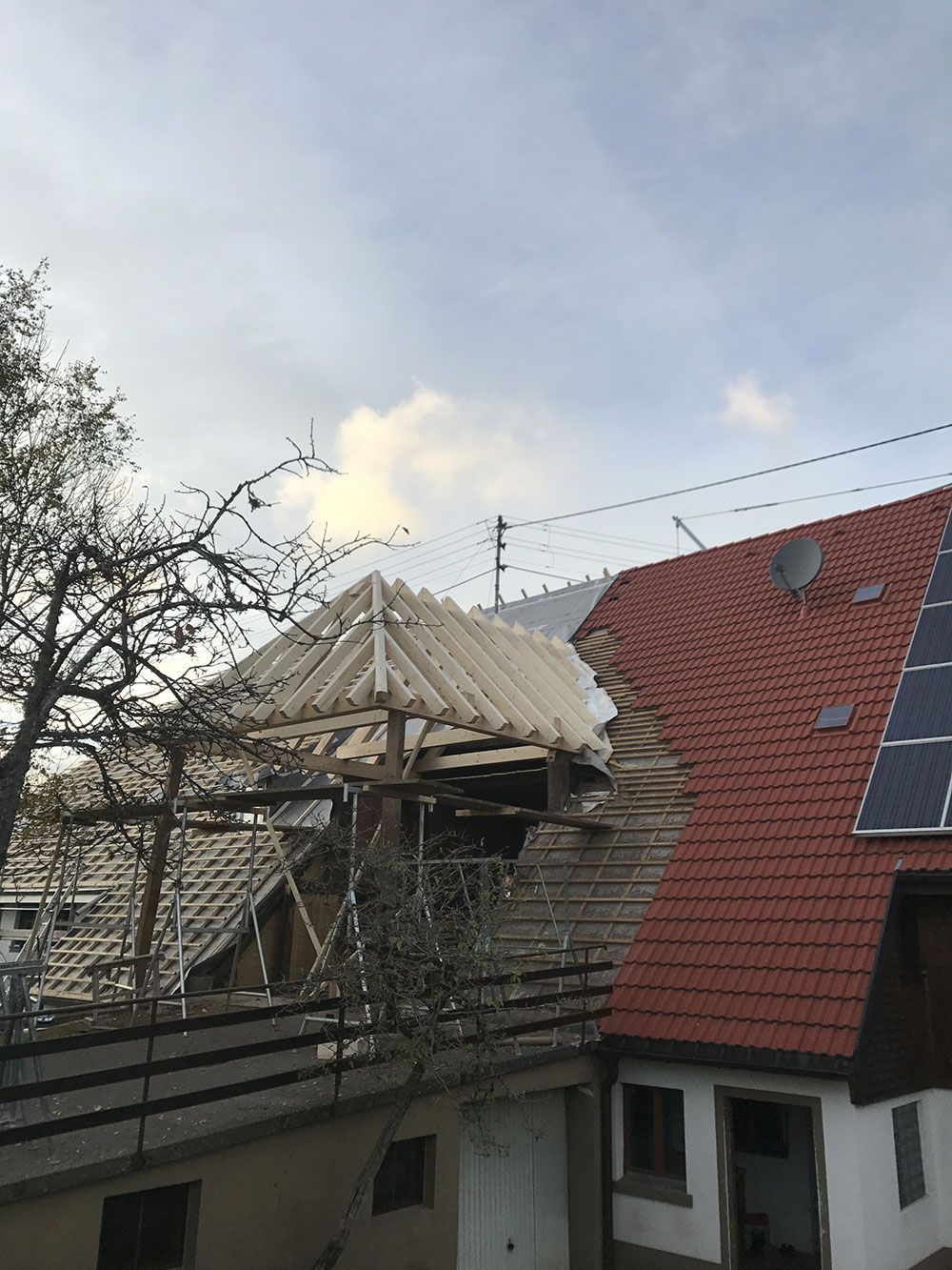 Ein im Bau befindliches hölzernes Dachgerüst neben einem bestehenden roten Ziegeldach mit Solarpaneelen an einem sonnigen Tag.
