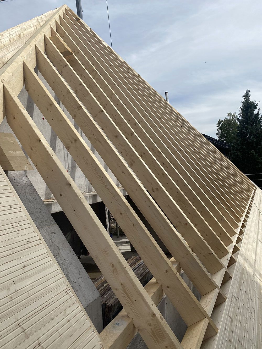 Eine Ansicht aus der Froschperspektive auf ein im Bau befindliches hölzernes Dachgerüst mit freiliegenden Sparren und Balken vor einem hellen, bewölkten Himmel.