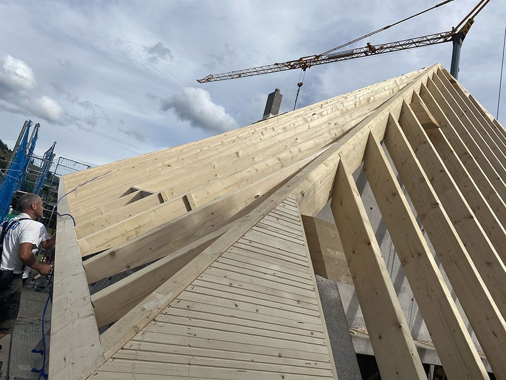 Ein Bauarbeiter errichtet unter einem teilweise bewölkten Himmel ein hölzernes Dachgerüst an einem Haus.