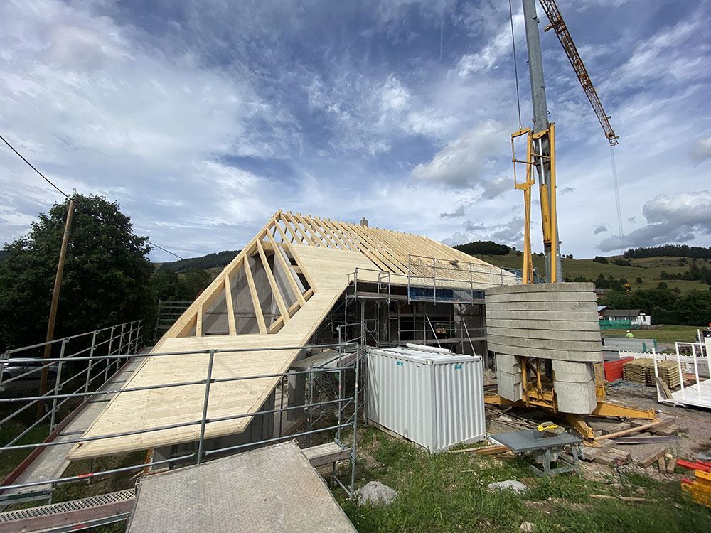 Baustelle mit einem Haus mit freiliegenden hölzernen Dachbalken und einem Kran unter blauem, bewölktem Himmel.