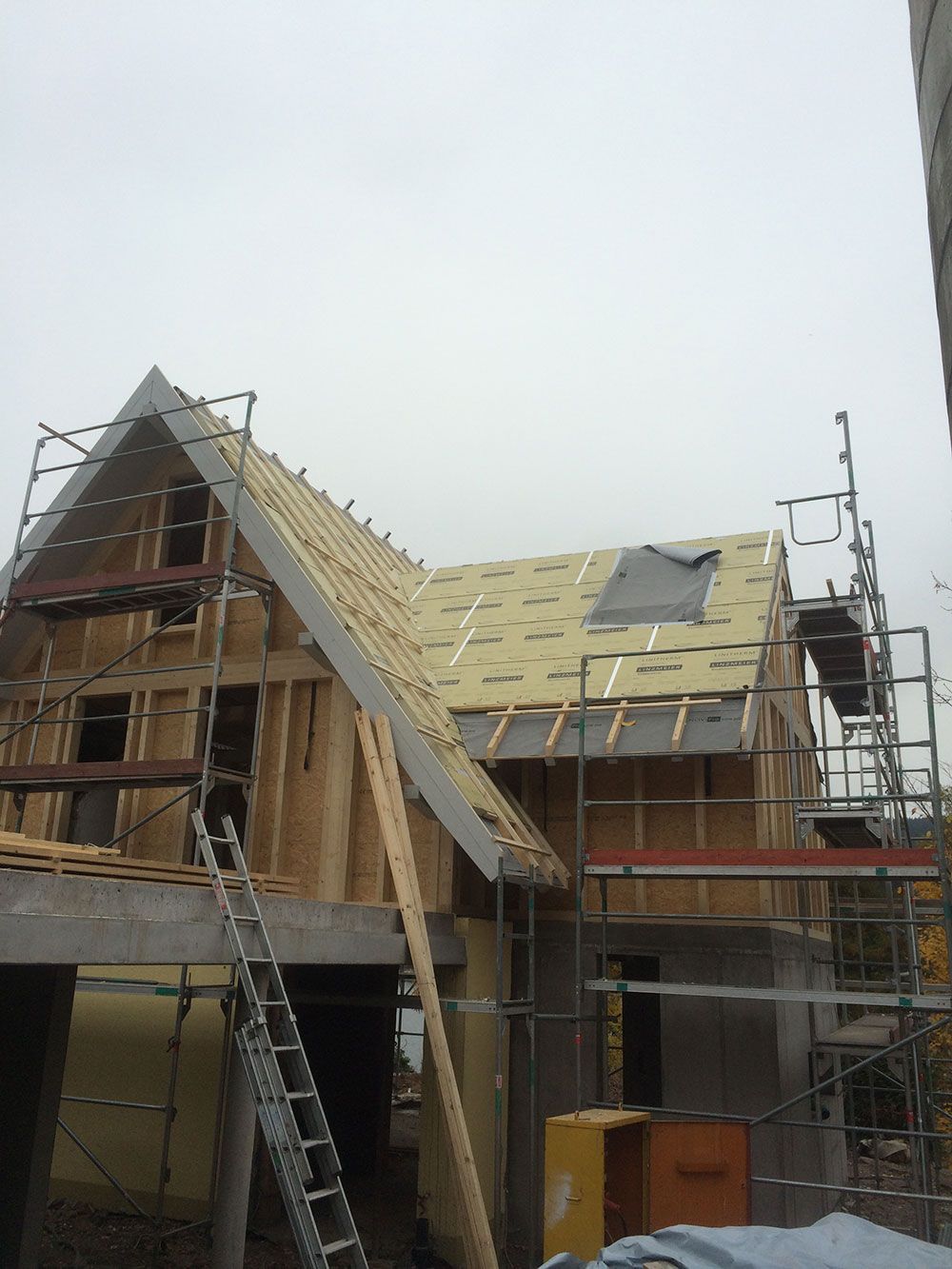 Ein im Bau befindliches Haus in Holzrahmenbauweise, teilweise mit Dämmplatten gedeckt und von einem Gerüst umgeben.