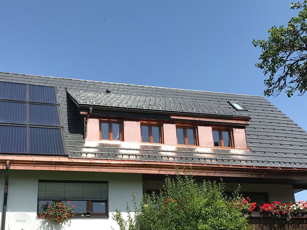Ein Hausdach mit dunklen Schindeln, einer Reihe von Solarmodulen und einer Dachgaube mit vier Holzfenstern.