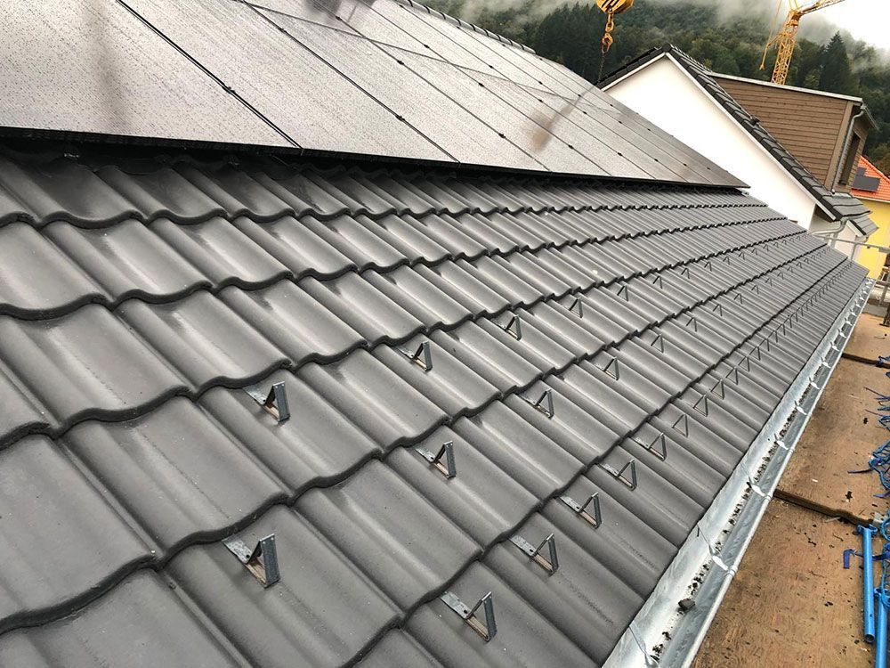 Ein schräges, graues Ziegeldach mit Schneefanggittern aus Metall und einer dunklen Solarmodulanordnung im oberen Bereich.