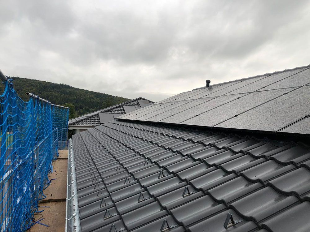 Blick auf ein dunkel geziegeltes Dach mit Solarpaneelen und blauem Sicherheitsnetz an der Seite unter bewölktem Himmel.