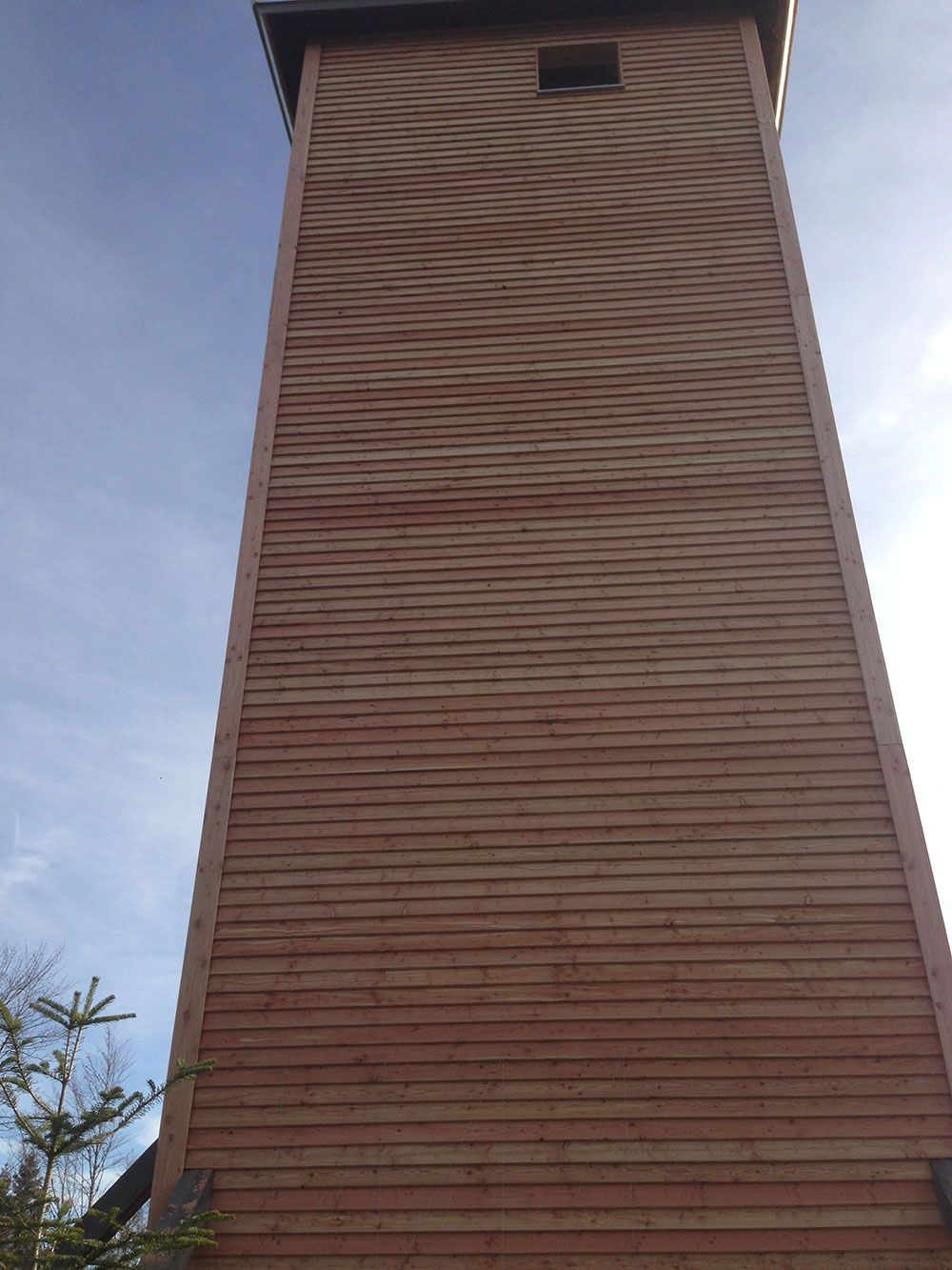 Ein hoher, rechteckiger Holzturm mit horizontalen Latten und einem kleinen quadratischen Fenster nahe der Spitze vor blauem Himmel.