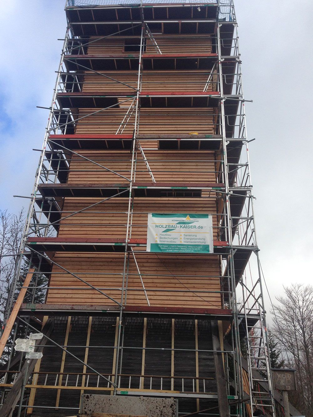 Ein im Bau befindlicher, mit einem Gerüst versehener Holzturm, an dessen Mittelteil ein Banner angebracht ist.