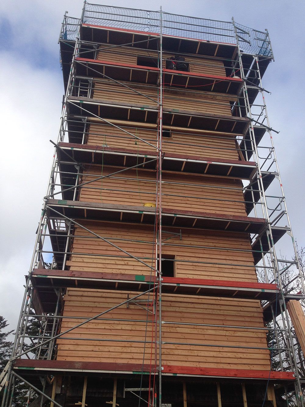 Ein im Bau befindlicher Holzturm, umgeben von grauen Metallgerüsten vor einem bewölkten Himmel.