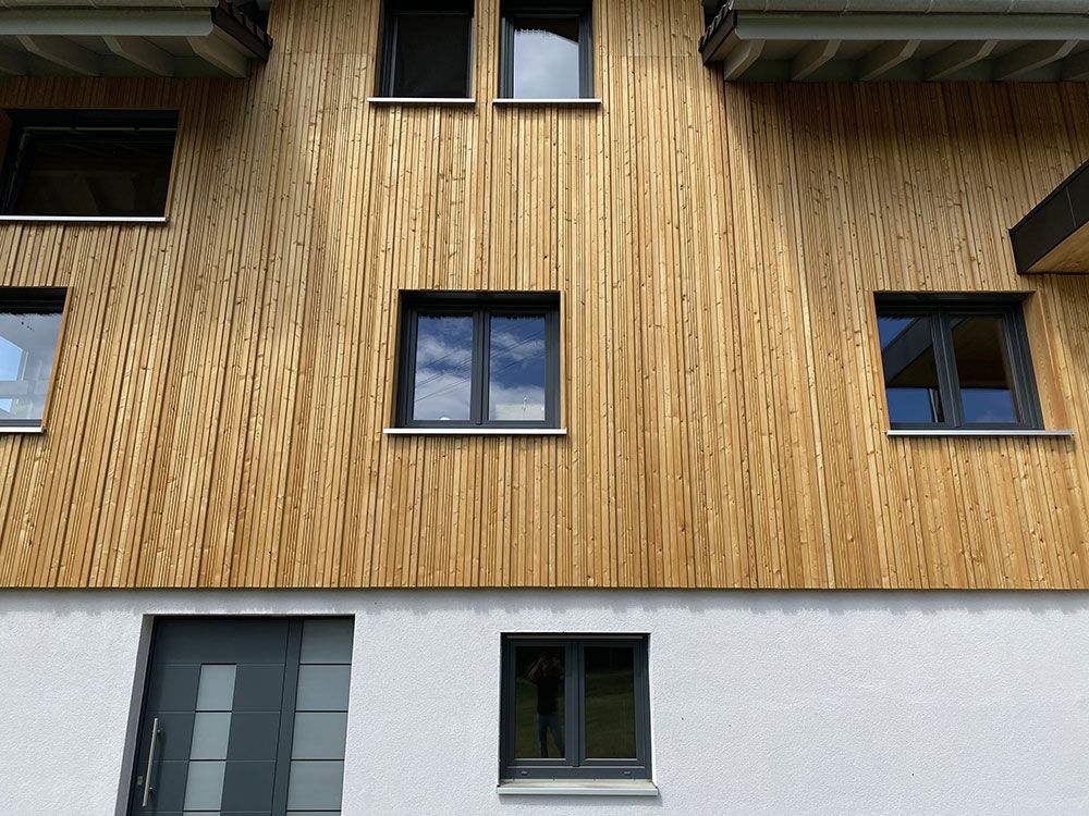 Ein modernes zweistöckiges Gebäude mit vertikaler, heller Holzverkleidung, einer grauen Tür und mehreren dunkel gerahmten Fenstern.