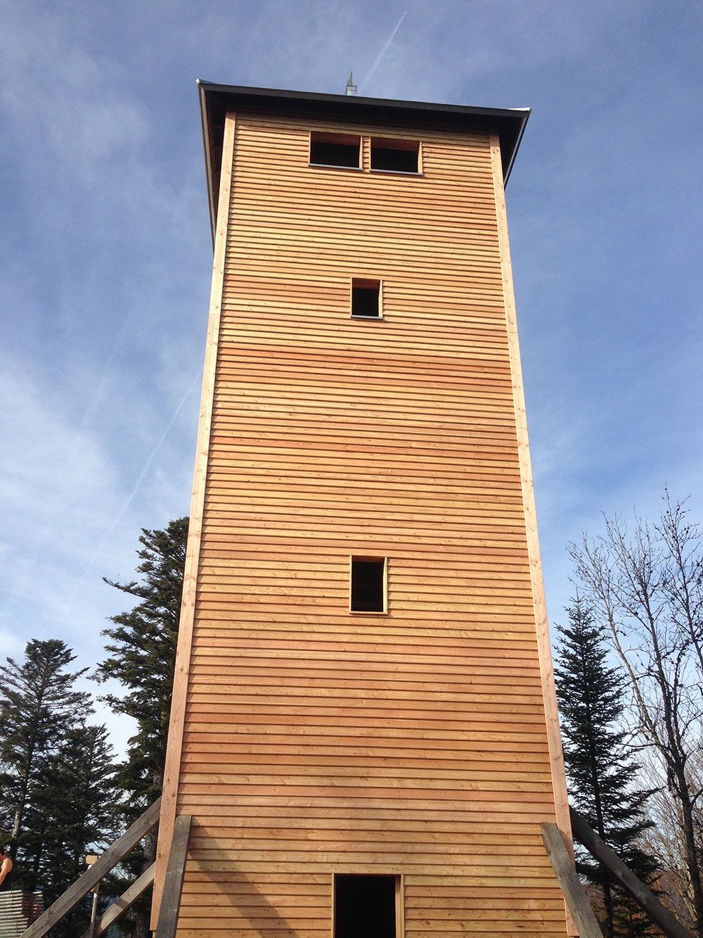 Ein hoher, hölzerner Aussichtsturm mit horizontalen Lamellen und rechteckigen Fenstern vor einem klaren blauen Himmel.