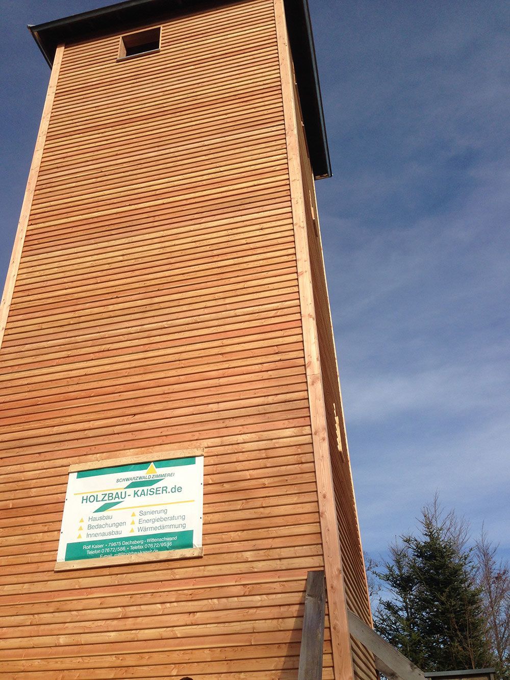 Ein hoher hölzerner Aussichtsturm vor blauem Himmel, an dessen Seite ein grün-weißes Schild angebracht ist.