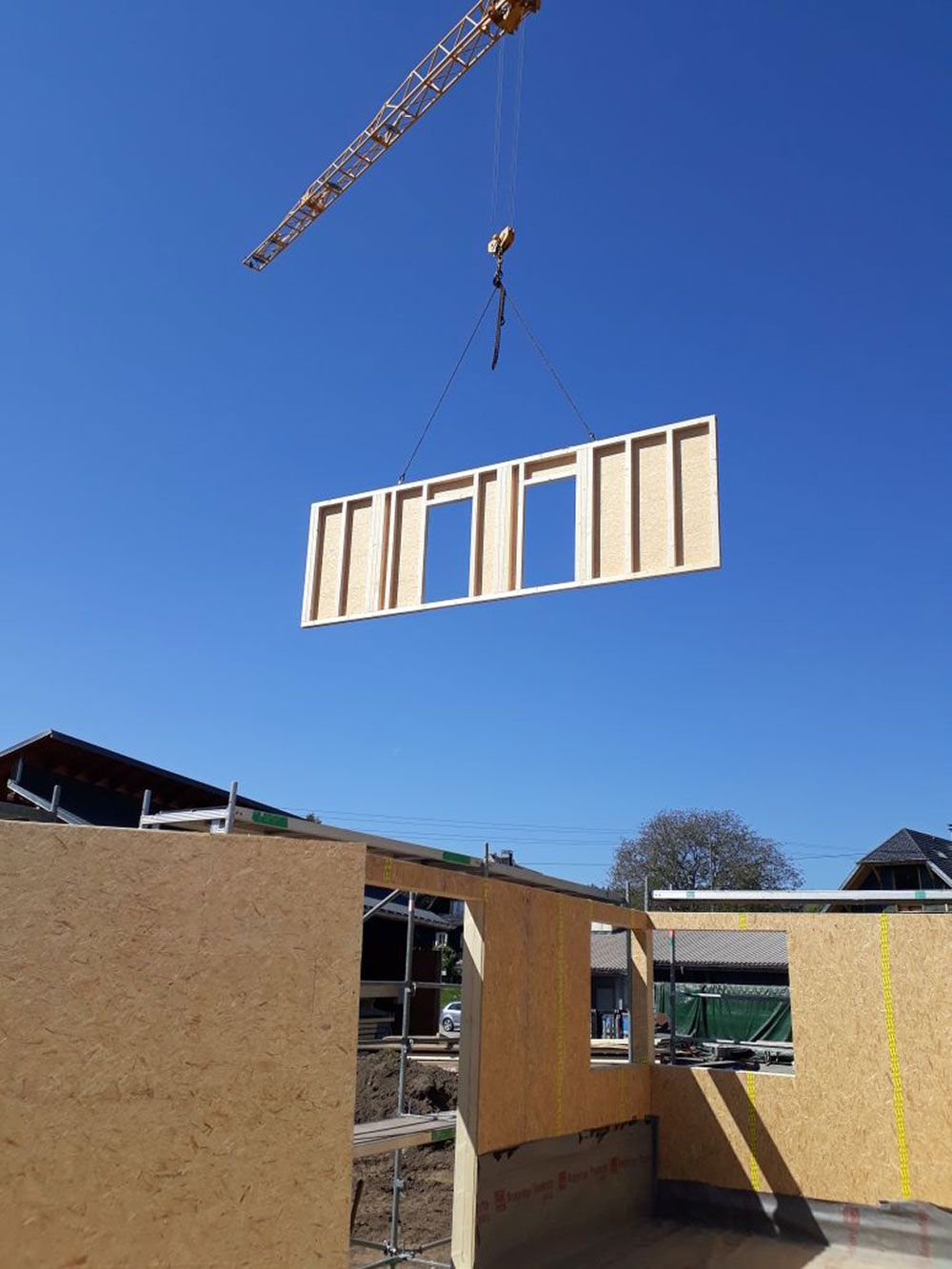 Unter strahlend blauem Himmel schwebt ein Baukran über einem Hausgerüst und hebt dabei eine vorgefertigte Holzwandplatte an.