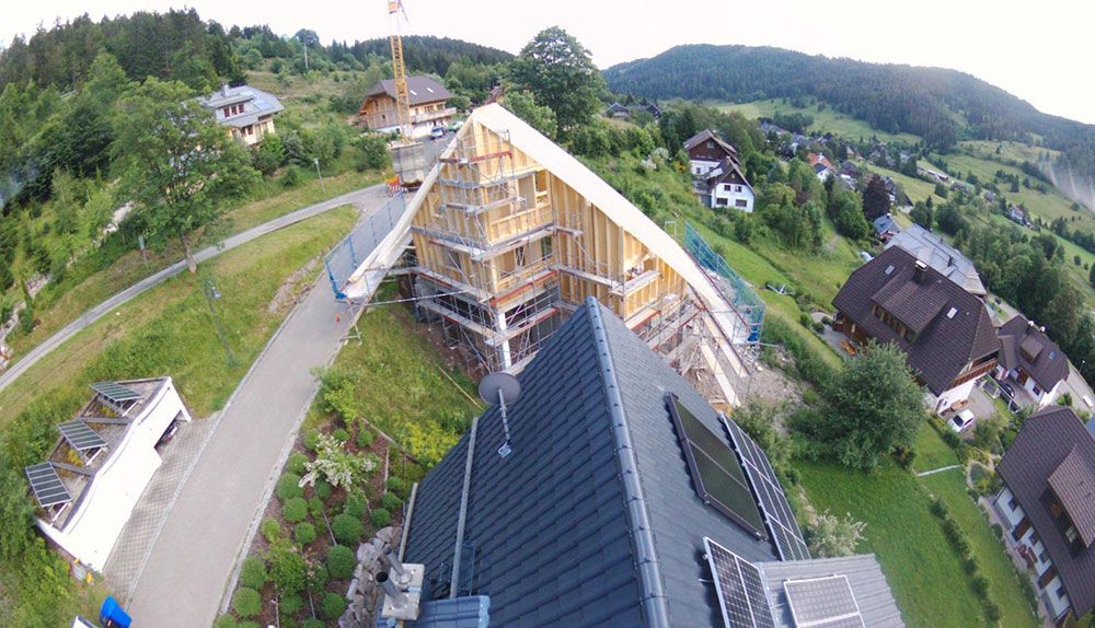 Luftaufnahme eines im Bau befindlichen Holzgerüsts an einem Hang, umgeben von Häusern, Straßen und grüner Landschaft.