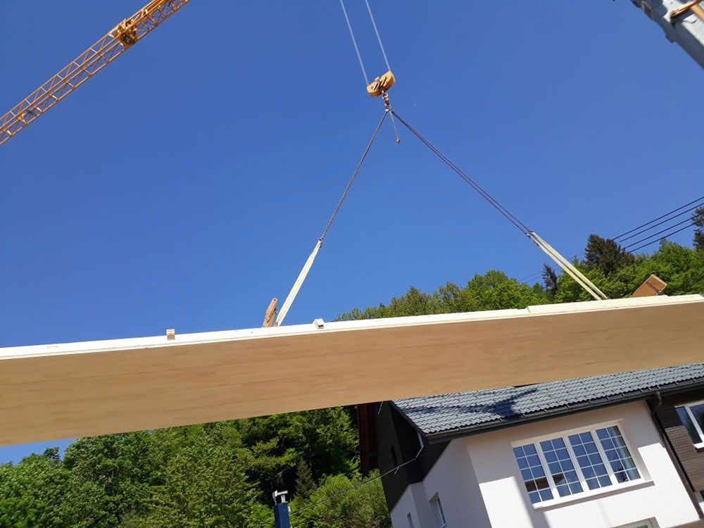 Ein Kran hebt einen langen Holzbalken vor einem klaren blauen Himmel in die Luft; im Hintergrund sind ein Haus und Bäume zu sehen.