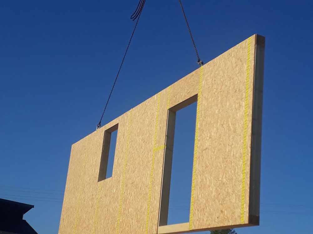 Ein Baukran hebt ein vorgefertigtes Wandpaneel aus OSB-Platten an, das zwei Fenster- oder Türausschnitte aufweist.