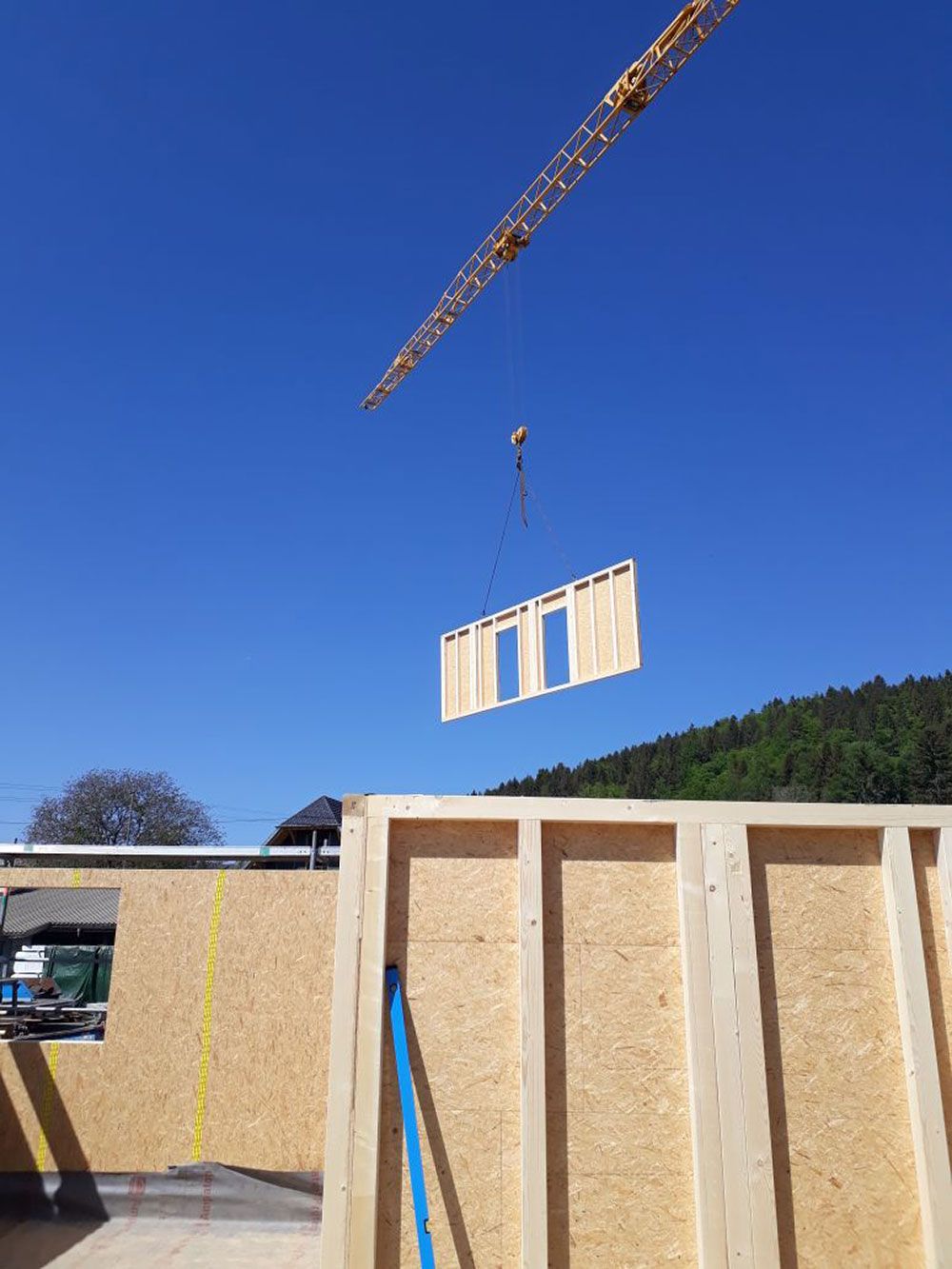 Unter strahlend blauem Himmel senkt ein Baukran ein vorgefertigtes Holzwandelement auf eine Baustelle.
