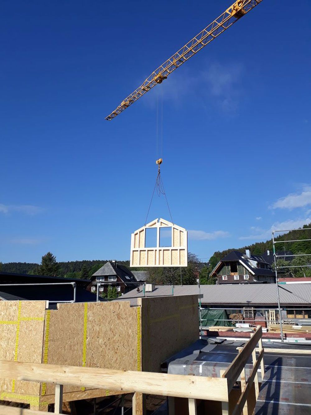 Ein Baukran hebt ein vorgefertigtes Holzwandelement unter strahlend blauem Himmel über eine Baustelle.