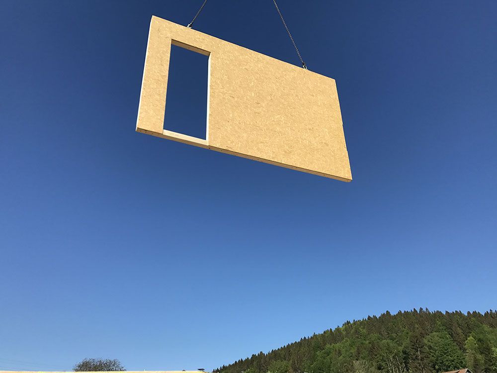 Eine vorgefertigte Holzwandplatte mit einem rechteckigen Türausschnitt wird vor strahlend blauem Himmel von einem Kran angehoben.