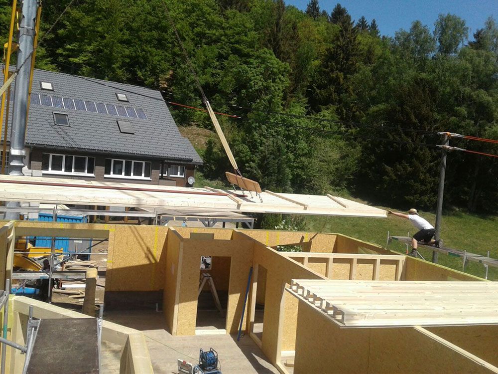 An einem sonnigen Tag senkt ein Kran eine große hölzerne Deckenplatte auf die Rahmenwände eines im Bau befindlichen Hauses.
