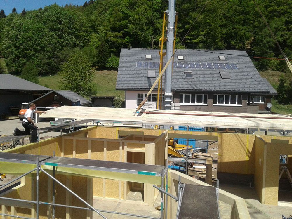 Eine Baustelle, auf der in der Nähe eines Hauses mit einem Kran Holzwandpaneele von einem Gerüst umgeben werden, die an ihren Platz herabgelassen werden.