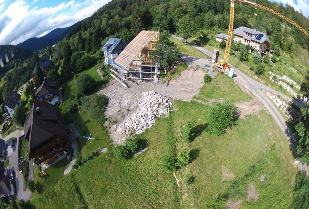 Luftaufnahme einer Wohnbaustelle mit einem Kran und einem im Bau befindlichen Holzhaus, umgeben von Grün.