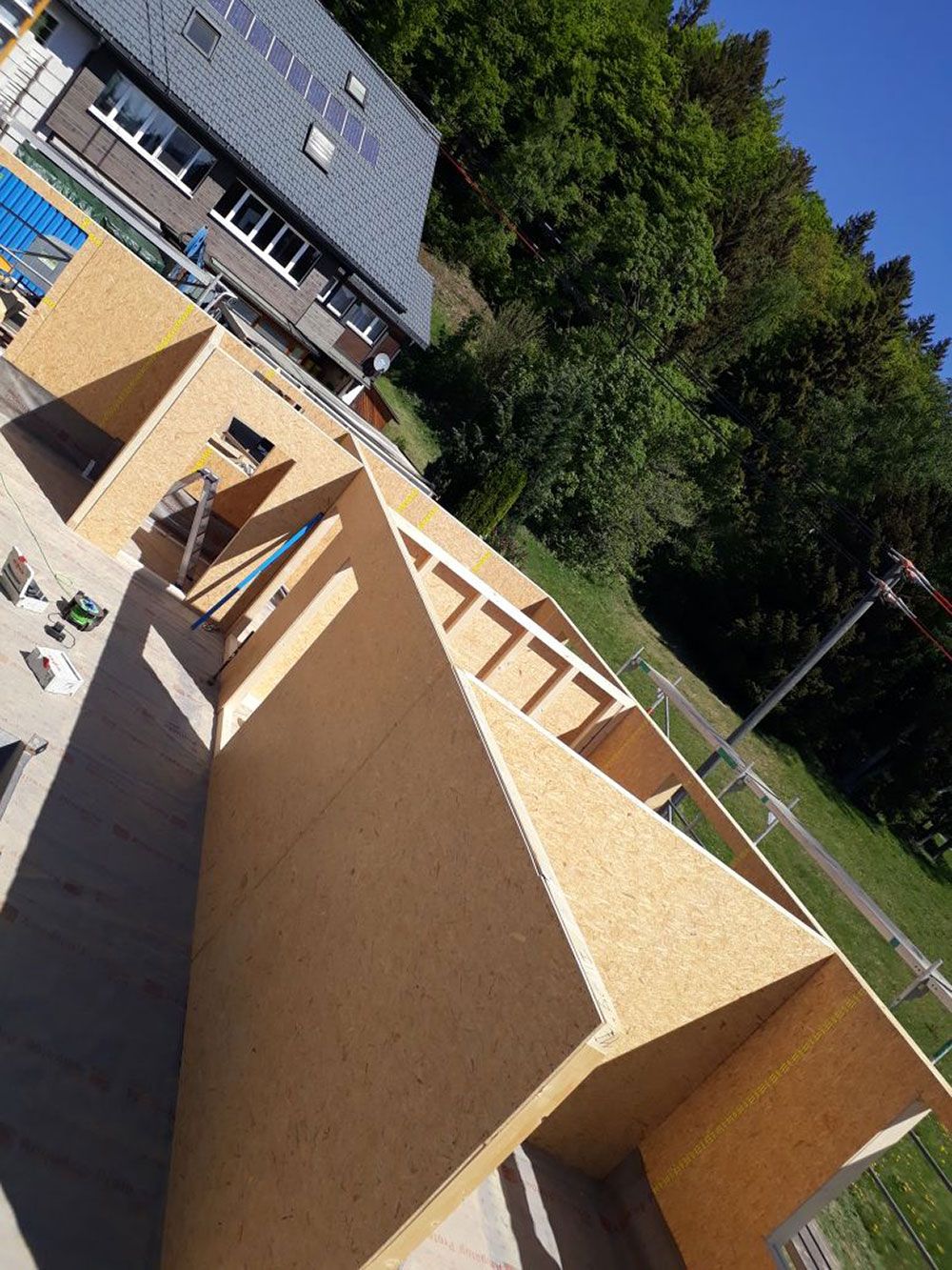 Ein Kran hebt eine große hölzerne Deckenplatte auf das Gerüst eines im Bau befindlichen Hauses in der Nähe eines Waldes.
