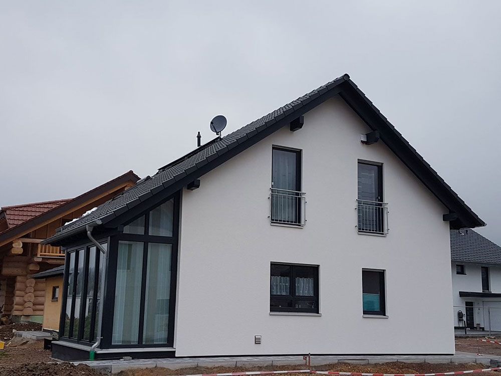 Ein modernes weißes Haus mit dunklem Giebeldach und schwarz gerahmten Fenstern, gelegen auf einem Grundstück unter bedecktem Himmel.