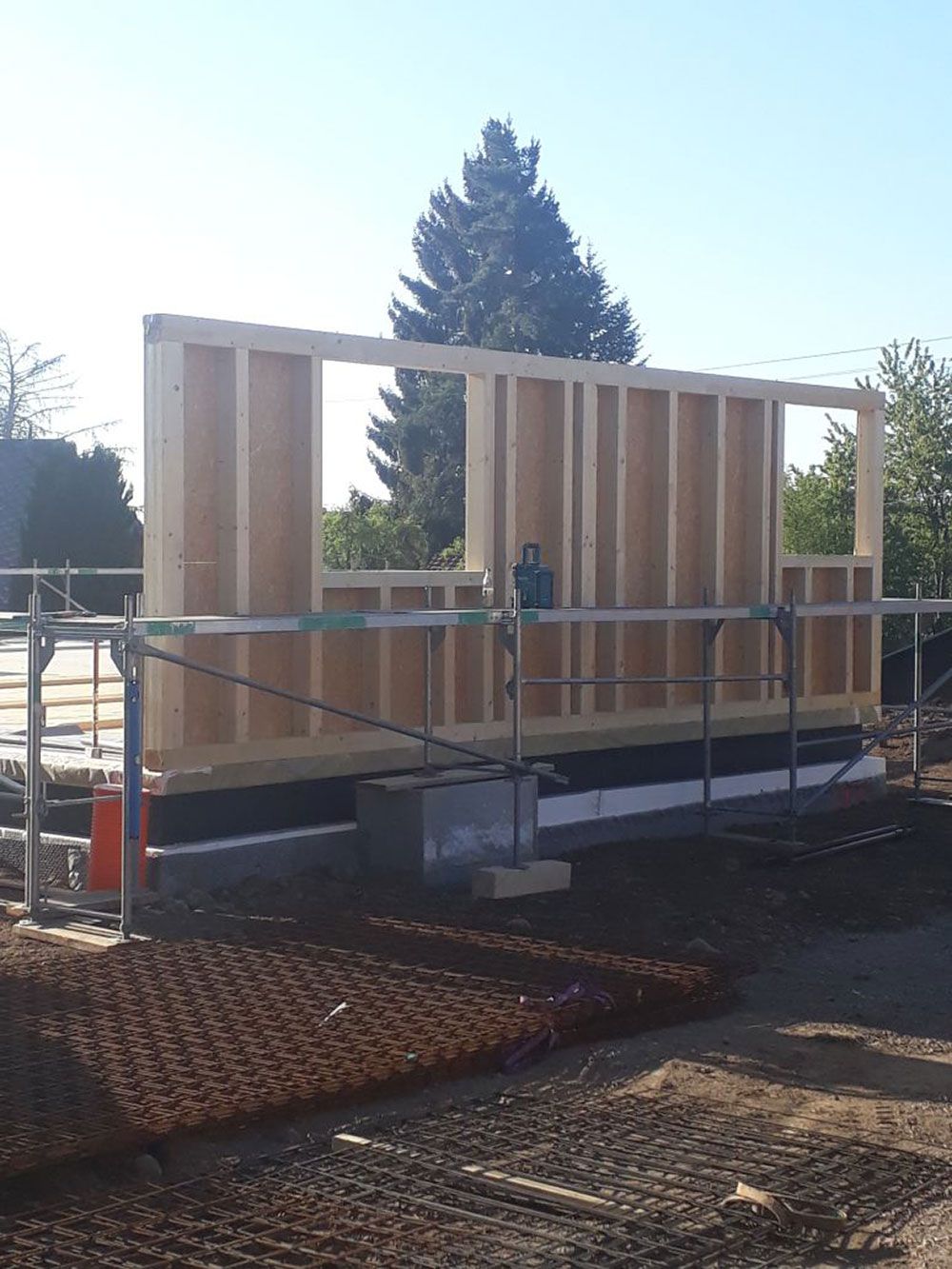 Eine im Bau befindliche Holzrahmenkonstruktion auf einem Betonfundament mit einer großen Fensteröffnung vor klarem Himmel.