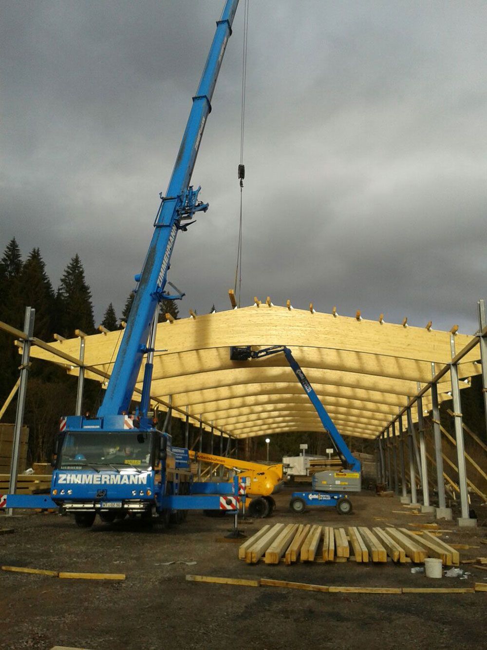 Unter bewölktem Himmel hebt ein großer blauer Kran einen gebogenen hölzernen Dachbalken auf eine Baustelle.