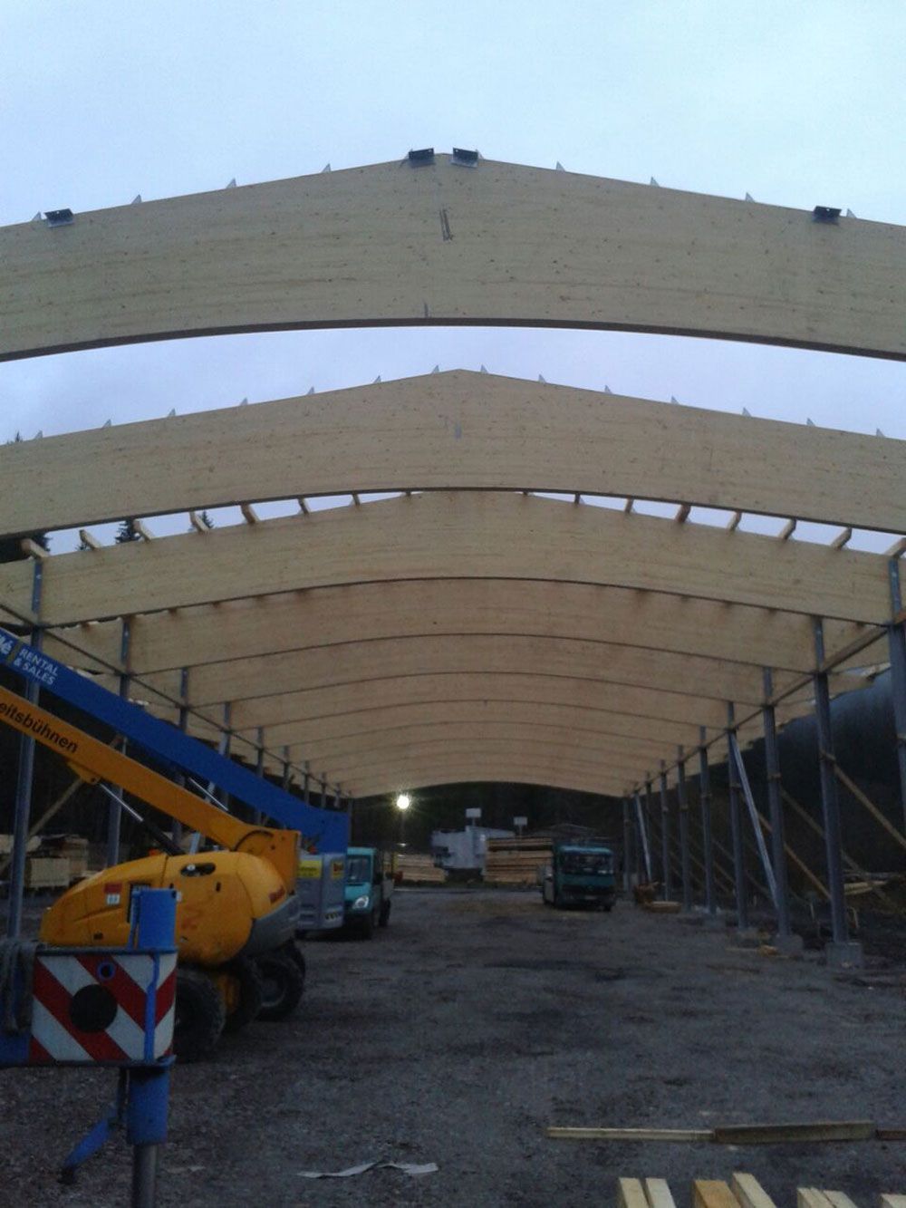 Die hölzerne Dachkonstruktion eines großen, im Bau befindlichen Industriegebäudes, auf dessen Baustelle sich schwere Maschinen befinden.