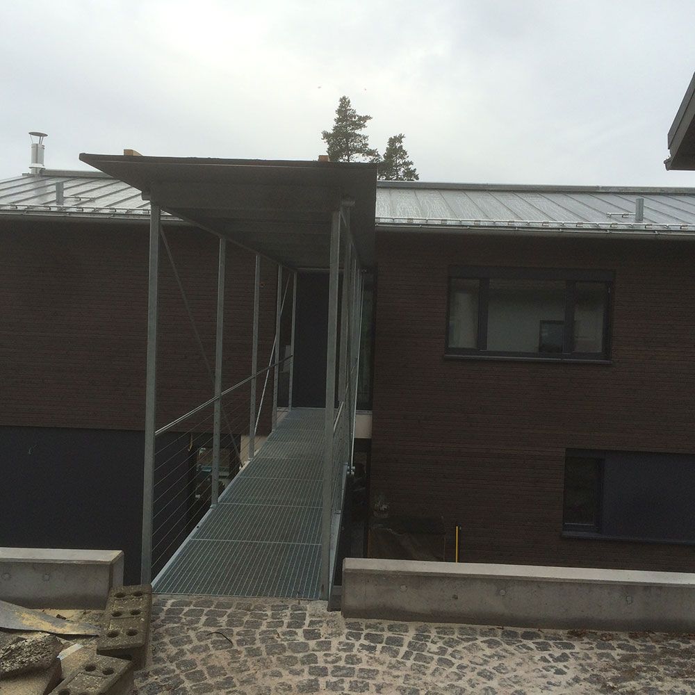 Ein metallener Steg mit Überdachung führt zum Eingang eines modernen, dunkelziegeligen Hauses mit Metalldach.