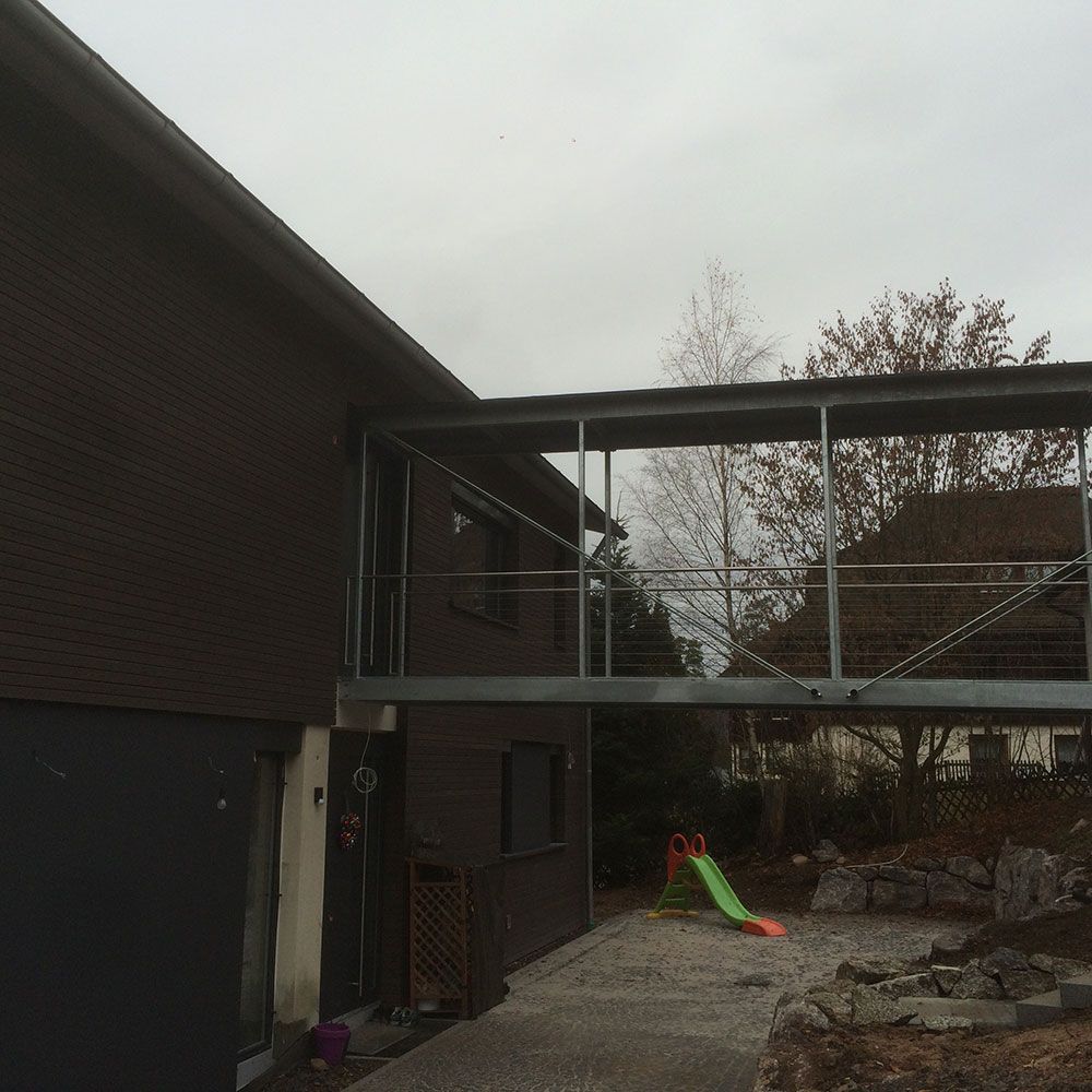 Ein Metallsteg verbindet das zweite Stockwerk eines braunen Hauses mit einem höher gelegenen Hofbereich, unter dem eine kleine Rutsche zu sehen ist.