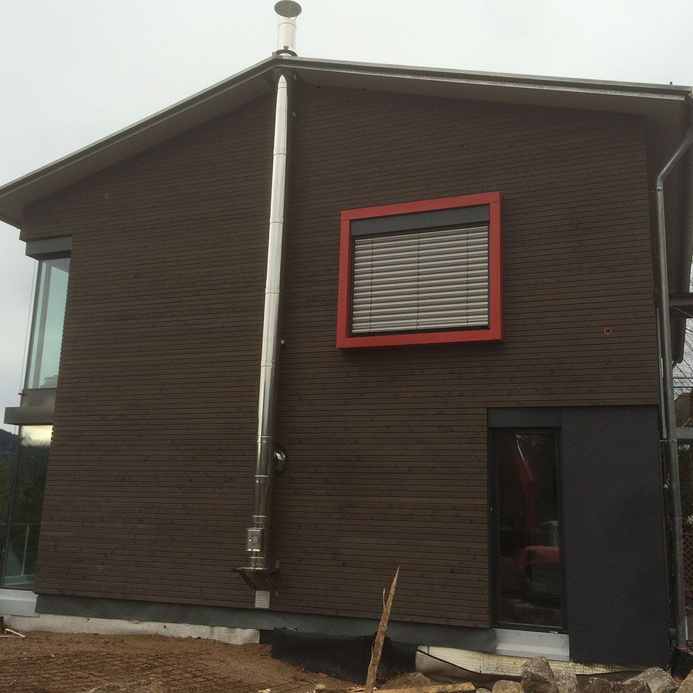 Ein dunkelbraunes Haus mit horizontaler Holzverkleidung, einem silbernen Schornstein, einem rot gerahmten Fenster und einer Glastür.