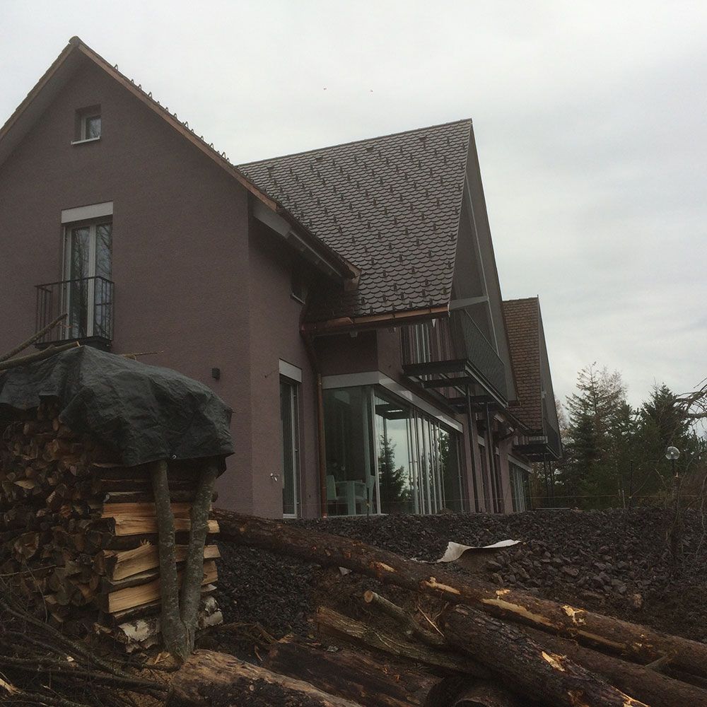 Ein zweistöckiges braunes Haus mit einem großen verglasten Wintergarten, im Vordergrund ein Haufen gestapeltes Brennholz und Baumstämme.