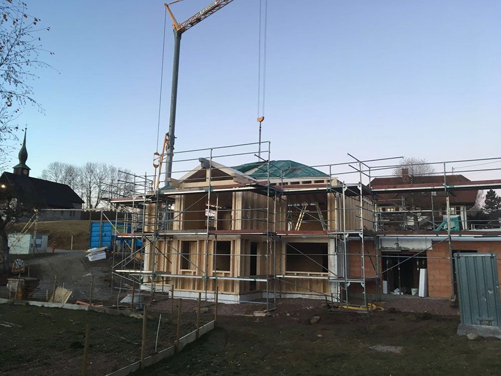 Ein zweistöckiges Holzhaus im Bau, umgeben von einem Gerüst, über dem ein Kran positioniert ist.
