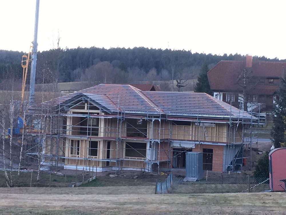 Ein zweistöckiges Haus im Bau mit Holzrahmenkonstruktion, Gerüst und Ziegeldach in ländlicher Umgebung.