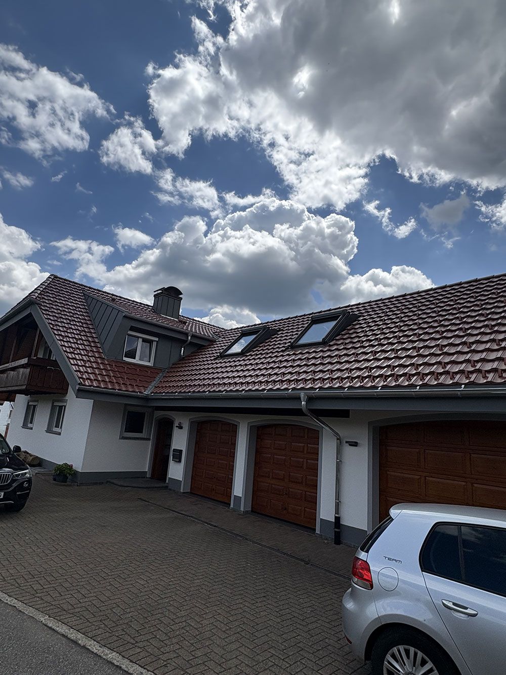 Ein zweistöckiges weißes Haus mit rotem Ziegeldach, drei Garagentoren aus Holz und zwei Dachfenstern unter einem bewölkten blauen Himmel.