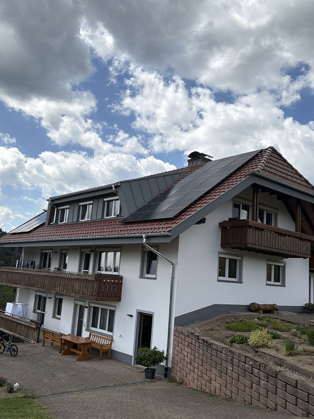 Ein mehrstöckiges weißes Haus mit dunklem Ziegeldach, Holzbalkonen und einer steinernen Stützmauer unter bewölktem Himmel.